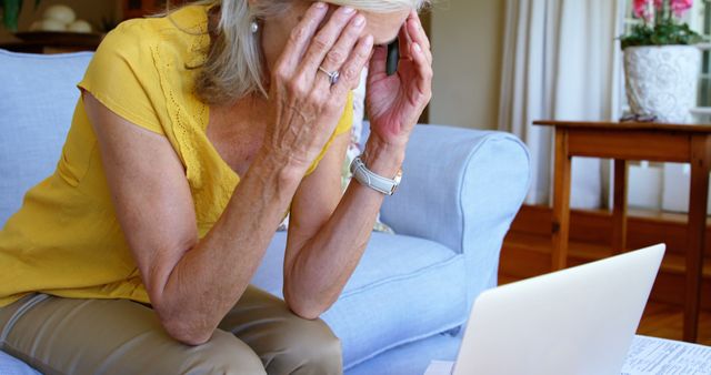 Senior woman wears yellow top, appears stressed while using a laptop in living room. Potential use for articles on elderly and technology, digital literacy, stress management, mental health, aging population adapting to modern life, or lifestyle choices for seniors.