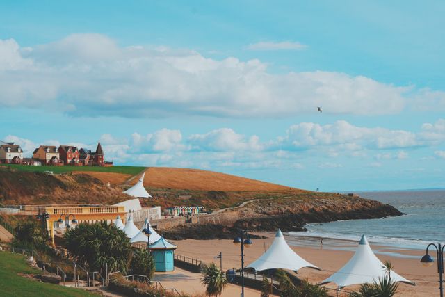 Coastal town featuring sandy beach, pavilions, and backdrop of houses under blue sky with clouds. Ideal for tourism, travel, vacation, and summer promotional materials. Perfect for illustrating seaside destinations, coastal scenery, and outdoor activities.