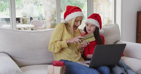 Mother and daughter opening christmas gift on sofa with laptop and santa hats