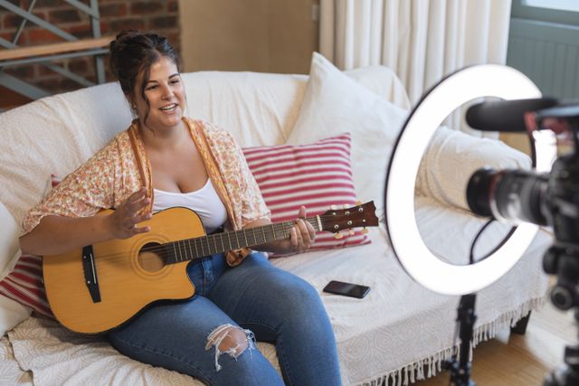 Caucasian female vlogger at home, holding guitar and talking during her online blog. Social distancing in quarantine lockdown during coronavirus covid 19 epidemic.
