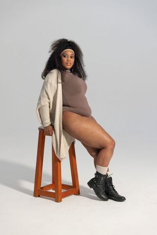 Curvy african american woman sitting on stool wearing brown bodysuit and cream cardigan