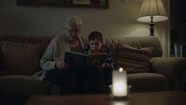 Grandmother reading bedtime story to grandson on cozy sofa by lamp and candlelight