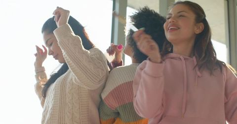 Young women dancing and laughing in sunlit room wearing sweaters and hoodie holding makeup