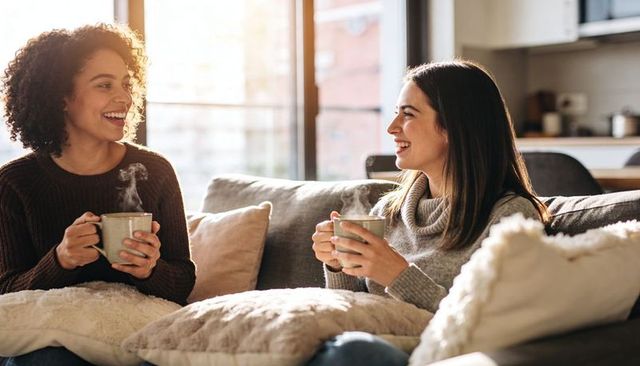 Two women sharing steaming mugs on cozy sofa, laughing and chatting in sunlit apartment