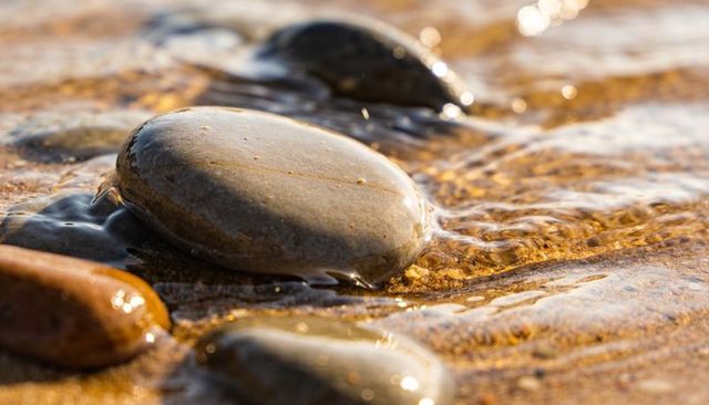 Glowing wet pebble reflecting warm sunlight on sandy shoreline with rippling water