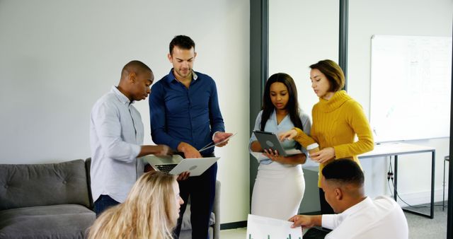 Professionals in an office collaborating on a business strategy. One person is holding a laptop while others discuss documents and graphs, creating a dynamic and productive atmosphere. Ideal for illustrating teamwork, business planning, modern work environments, diverse workforces, and strategy sessions.