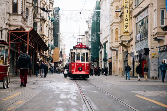 Vintage red tram traveling through busy European city street with pedestrians walking along both sides. The area features historic architecture and various shops, creating a vibrant urban scene. Useful for illustrating city life, tourism, public transportation, and historic urban areas.