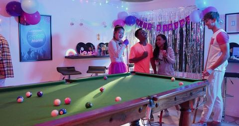 Young friends singing and playing billiards at birthday party with balloons and disco decor