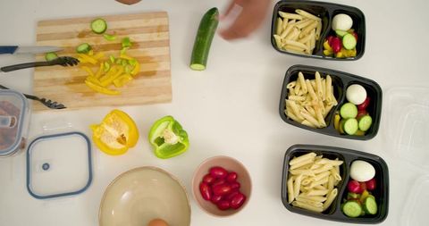 Packing three black meal-prep containers with penne, boiled egg, cucumber and tomatoes