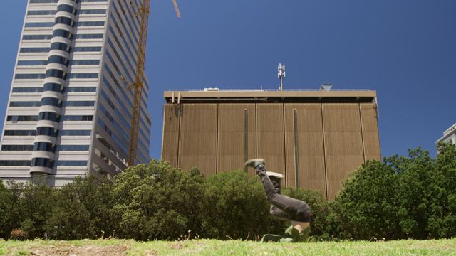 Caucasian man practicing parkour and performing acrobatics on grass near tall city buildings on a sunny day. Great for illustrating urban agility, fitness activities, athletic training, or energetic outdoor lifestyles.