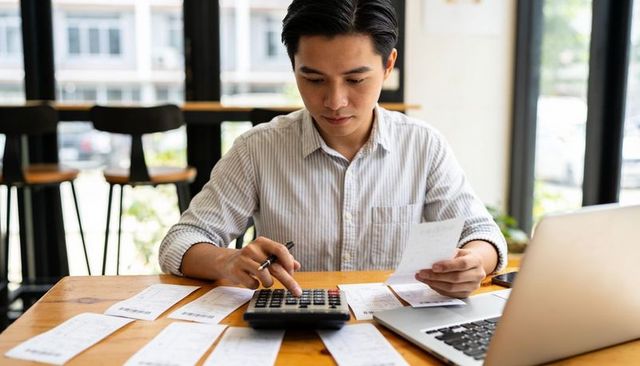 Man calculating personal finances with calculator and receipts at laptop workspace in daylight