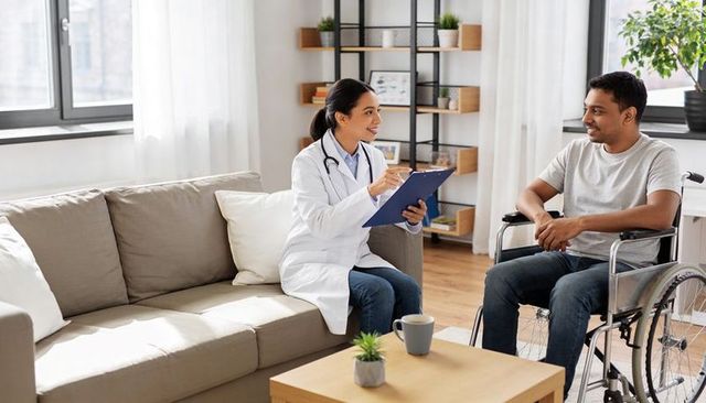 Female doctor consulting male wheelchair patient during home visit in modern living room