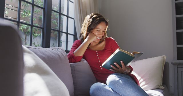 Woman is reading a book and smiling on a cozy couch near a window, with sunlight streaming in. Ideal for use in lifestyle blogs, advertisements for home decor, relaxation, or leisure activities, and promoting books or reading.