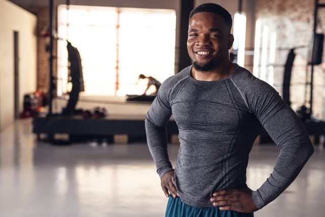 Young African American male boxer standing confidently in a gym with a smile on his face. He is wearing sportswear and has his hands on his hips. This image can be used for promoting fitness, boxing training programs, health clubs, and motivational content related to sports and exercise.