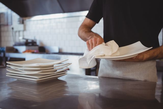 Waiter cleaning plates in a restaurant kitchen, ensuring hygiene and cleanliness. Ideal for use in articles or advertisements related to restaurant operations, food service, hospitality industry, and cleanliness standards.