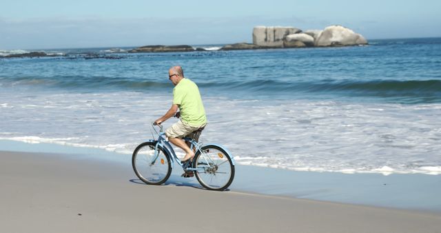 Senior man casually biking on a sandy beach along the ocean waves, embracing nature and enjoying an active lifestyle. Ideal for ads about healthy living, outdoor activities, leisure, vacations, retirement, and exercise habits. Can also be used in brochures, wellness campaigns, and travel promotions.