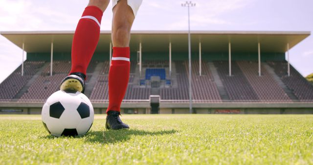 Image of a football player wearing red socks dribbling a soccer ball on a grassy field in an empty stadium. This can be used for sports-related promotions, fitness campaigns, or soccer training materials.