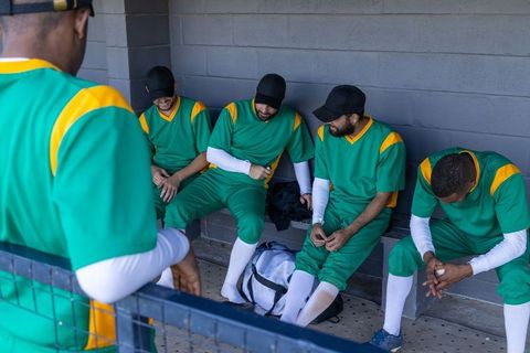 Baseball team waiting in dugout wearing green uniforms, players relaxing on bench