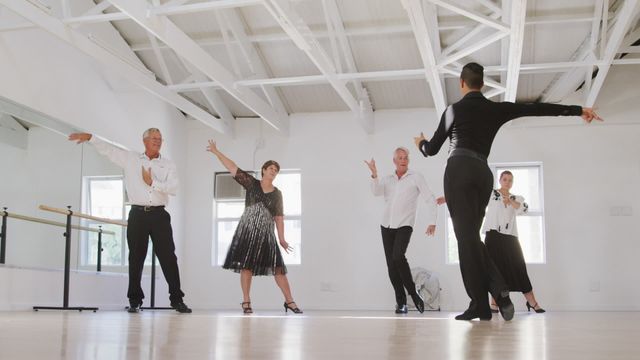 A biracial male dance teacher demonstrating dance steps to a group of Caucasian seniors in a bright dance studio. The elegant studio is marked by white walls and a well-lit environment with reflections from large windows. The seniors, dressed smartly, follow the instruction with keen interest. This video is useful for promoting dance classes, instructional content, senior activities, healthy lifestyles, and showcasing dance as a beautiful form of fitness and community engagement.