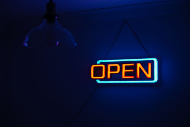 Bright neon open sign hanging on a dark wall, with a vibrant blue and red glow. Ideal for illustrating business hours, advertising, marketing, and creating a retro or urban atmosphere. Suitable for marketing agencies, social media banners, business advertisements, and website headers.