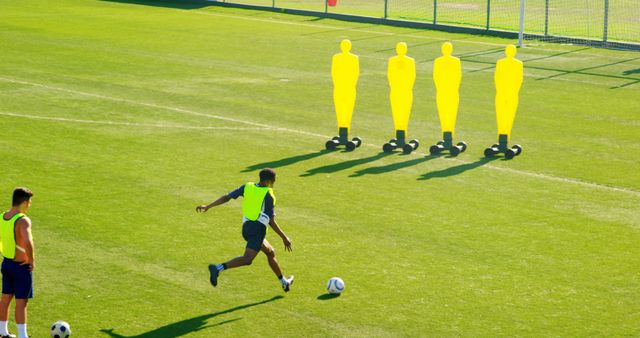 Soccer players practicing on a football field with bright yellow training dummies. One player is captured in motion, attempting to kick the ball. Suitable for illustrating articles about soccer training, teamwork in sports, athletic drills, and outdoor exercises.