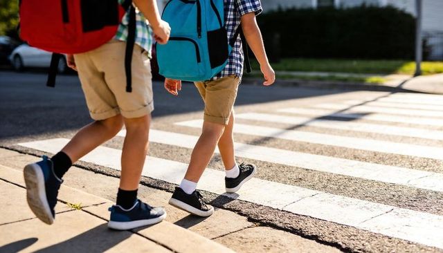 Two students crossing sunlit suburban crosswalk wearing colorful backpacks