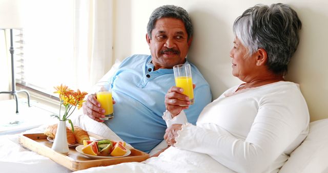 Senior couple comfortably enjoying breakfast in bed. Both are holding glasses of orange juice while smiling and conversing. Tray with croissants, fruit, and flowers adds to the cozy atmosphere. Great for health and wellness topics, senior living, retirement, and lifestyle blogs.