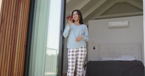 Smiling asian woman speaking into phone at bedroom doorway wearing cozy loungewear