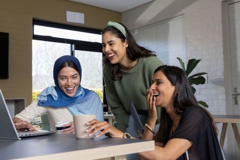 Diverse women laughing and collaborating around laptop in modern home office workspace