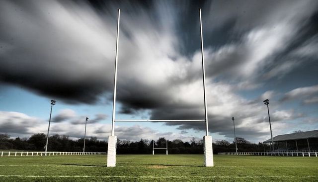Dramatic rugby goalposts on green pitch under dynamic cloudy sky with stadium lights