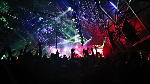 Image depicting an energetic crowd enjoying a live music concert with vibrant lighting. The audience has their hands in the air, showcasing excitement and enjoyment. This can be used to illustrate the lively and festive atmosphere of music festivals, concerts, and other large-scale live events.