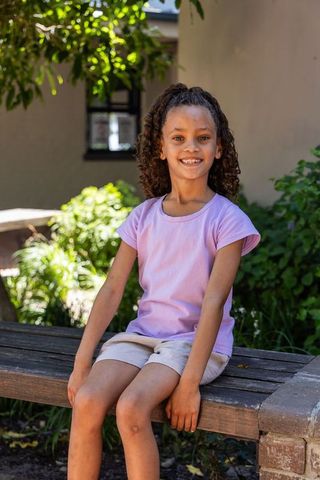 Sunlit smile: school-age girl sitting on garden bench wearing lilac shirt and shorts
