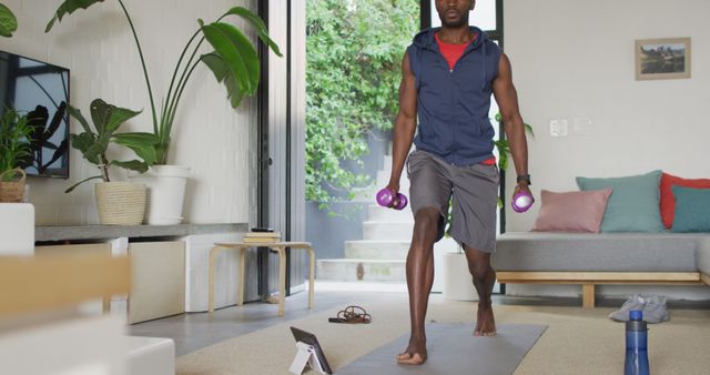 Young man performing a workout routine at home, using dumbbells for strengthening exercises. Living room features modern decor with indoor plants, a couch, and bright natural light. Ideal for illustrating topics related to home fitness, wellness, indoor activities, or online workout programs.