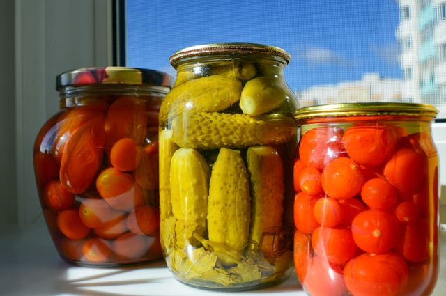 Assorted glass jars filled with pickled vegetables, including tomatoes and cucumbers, placed on a sunlit window sill. This vibrant and colorful composition is perfect for use in articles or blogs on homemade food preservation, healthy eating, and urban kitchens. It could also be useful in marketing materials for kitchen and pantry products or cooking magazines.