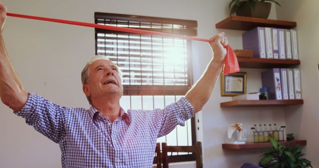 Senior man using resistance band for exercise in a cozy home office. Perfect for portraying active lifestyles, physical therapy routines, and healthy living habits for elderly individuals. Ideal for articles on fitness, rehabilitation, or senior well-being.