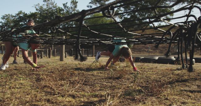 Group of individuals participating in an outdoor obstacle course, crawling under a rope net with determination and focus. Perfect for use in materials related to fitness, teamwork, and outdoor adventures. Can be used in advertisements for gyms, fitness classes, outdoor equipment, or team-building activities.