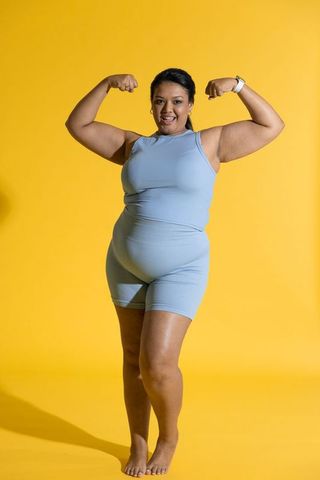 Mid-adult woman flexing arms wearing blue ribbed activewear and smartwatch on yellow background