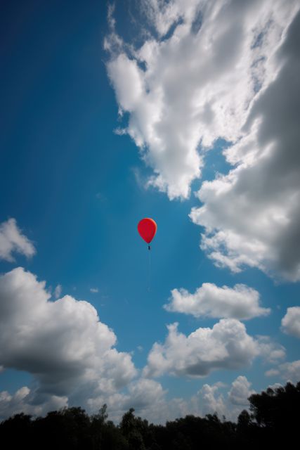Red balloon in cloudy blue sky over tree tops, copy space, created using generative ai technology. Freedom, celebration and independence, digitally generated image.