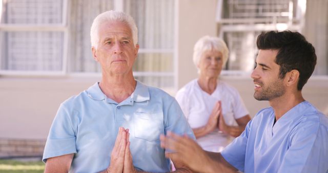 Senior man with white hair practicing meditation with hands in prayer position, guided by young caregiver in blue uniform, outdoors in a garden setting. Older woman in background also meditating. Ideal for promoting elderly care, wellness programs, physical therapy, mindfulness, and retirement living.