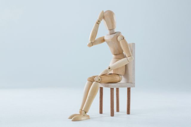 Wooden figurine sitting on a chair with hand on forehead, conveying a sense of tiredness and stress. Ideal for illustrating concepts of fatigue, mental health, stress, and contemplation. Suitable for use in articles, blogs, and educational materials about mental well-being and stress management.