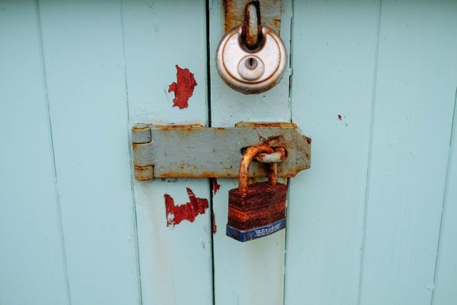 A close-up of a rusty padlock securing a weathered blue wooden door. The peeling paint and oxidation on the lock create a vintage and aged appearance. This image can be used to represent themes of security, locking mechanisms, age, decay, and rustic aesthetics.