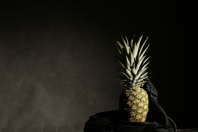 Pineapple sits on black cloth against dark backdrop, creating a strong contrast and focusing attention on fruit. Perfect for use in healthy eating promotions, tropical themed designs, or minimalist art. Emphasizes freshness and juiciness of pineapple, showcases simplicity and elegance.