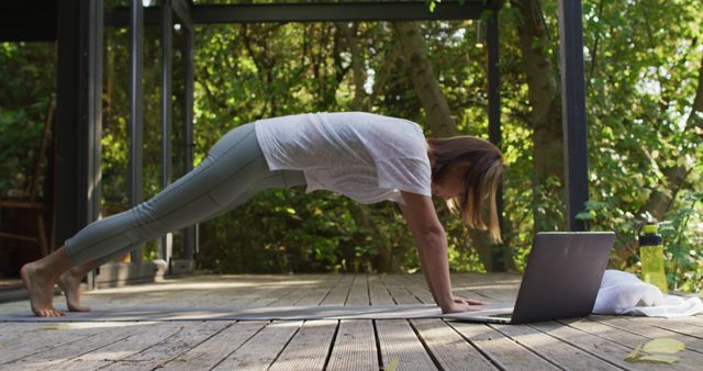 Woman practicing yoga during an online class in a serene outdoor environment surrounded by greenery. She is in a plank position, engaging in a fitness routine with a laptop for guidance. This image is excellent for promoting online fitness programs, outdoor workout gear, healthy lifestyles, and yoga equipment.