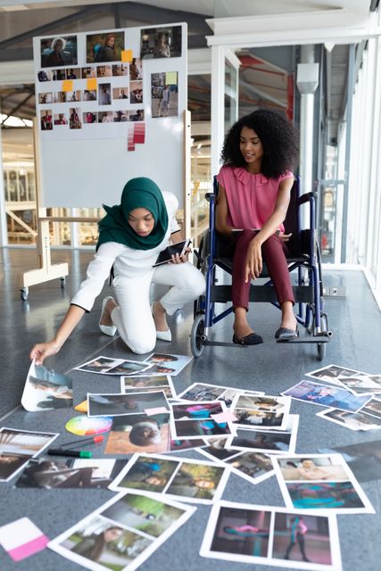 Two young female graphic designers are reviewing photographs spread out on the floor in a modern office. One woman is wearing a hijab and the other is in a wheelchair, highlighting diversity and inclusion. This image can be used to represent teamwork, creative collaboration, and diversity in the workplace, making it suitable for articles, blogs, and promotional materials related to business, design, and inclusivity.