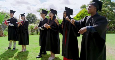 Diverse graduates celebrating on lawn wearing caps and gowns holding diploma scrolls