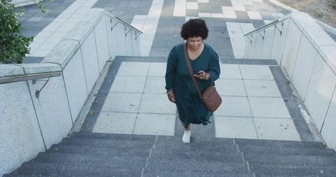 Woman ascending urban stairs while checking smartphone, wearing teal dress and crossbody bag