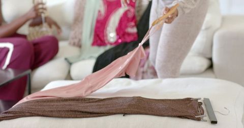 African american women comparing silky pink top and shimmery brown dress on cozy sofa