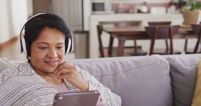 Senior woman smiling while using a digital tablet with headphones on cozy sofa in comfortable living room. Perfect for illustrating aging in place, technology usage by older adults, digital entertainment for seniors, home comfort, leisurely activities, and relaxing lifestyle.