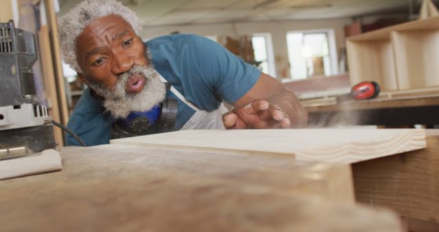 African american male carpenter blowing the dust over wooden plank at a carpentry shop. carpentry, craftsmanship and handwork concept