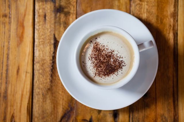 Overhead view of a cappuccino with foam and cocoa powder on top, placed on a rustic wooden table. Ideal for use in coffee shop promotions, breakfast menus, lifestyle blogs, and social media posts about cozy mornings and relaxation.